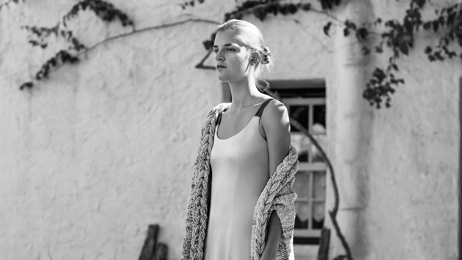 Model wearing a long open knitted wrap over a fluid dress in a black-and-white outdoor photograph