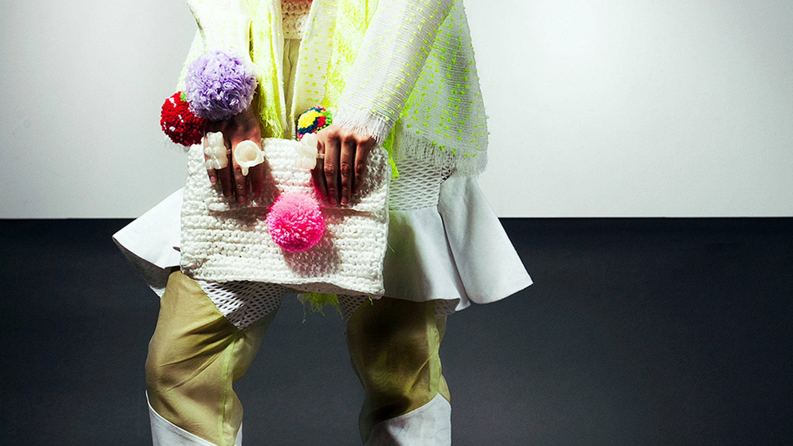 Hands holding a white knitted bag with vivid pink, red, lilac, and green pompom details