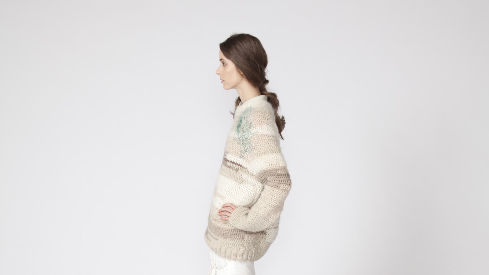 Side-profile model in an oversized neutral hand-knit sweater standing against a pale studio background