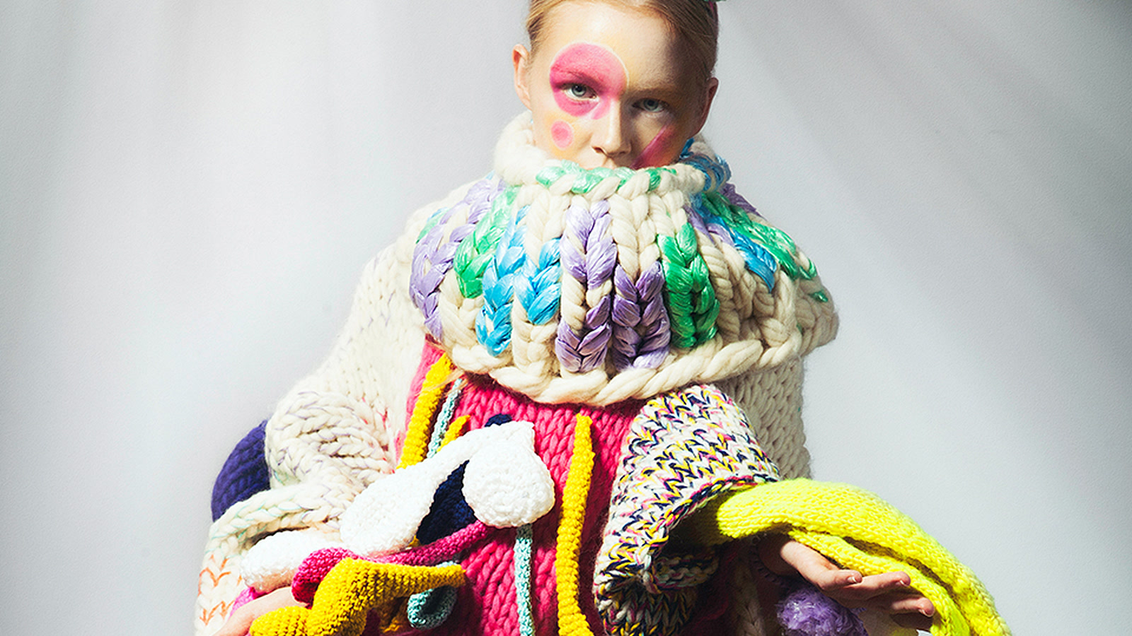 Model wearing a highly structured multicoloured knitted construction with an oversized raised collar and projecting tubular forms
