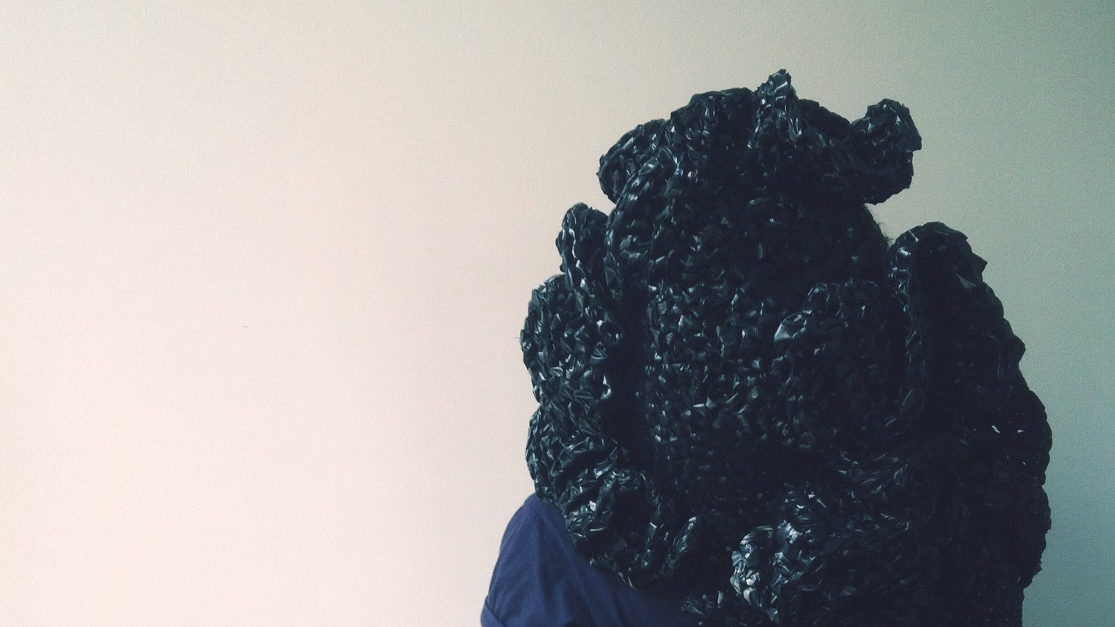 Person carrying a monumental black knitted structure against a pale studio wall