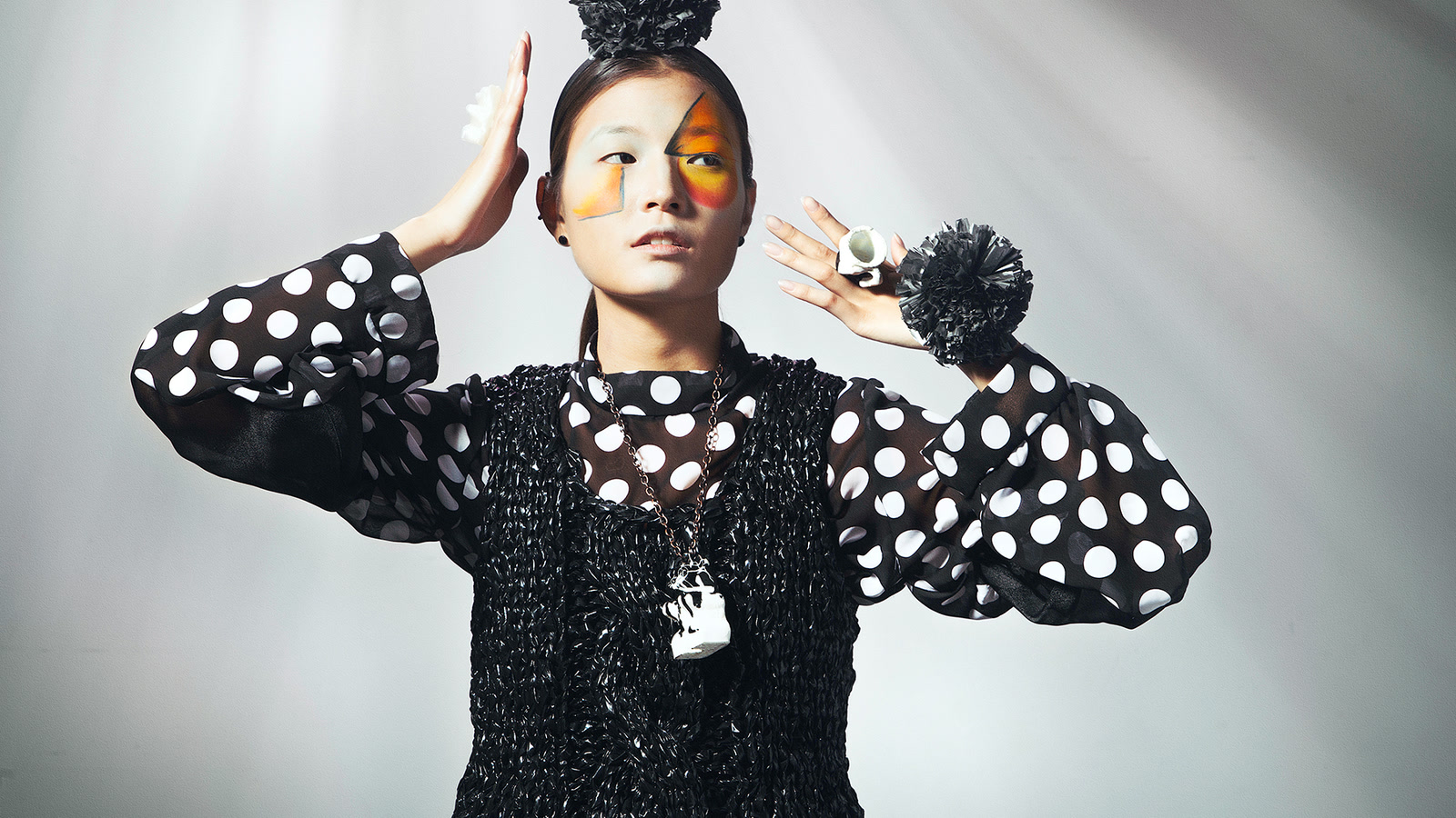 Model raising both arms while wearing a black open-loop knitted vest over a spotted blouse