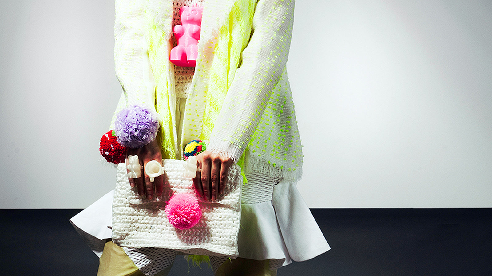 Close editorial view of a white knitted look with fluorescent yellow details, multicoloured pompons, and a textured knitted bag