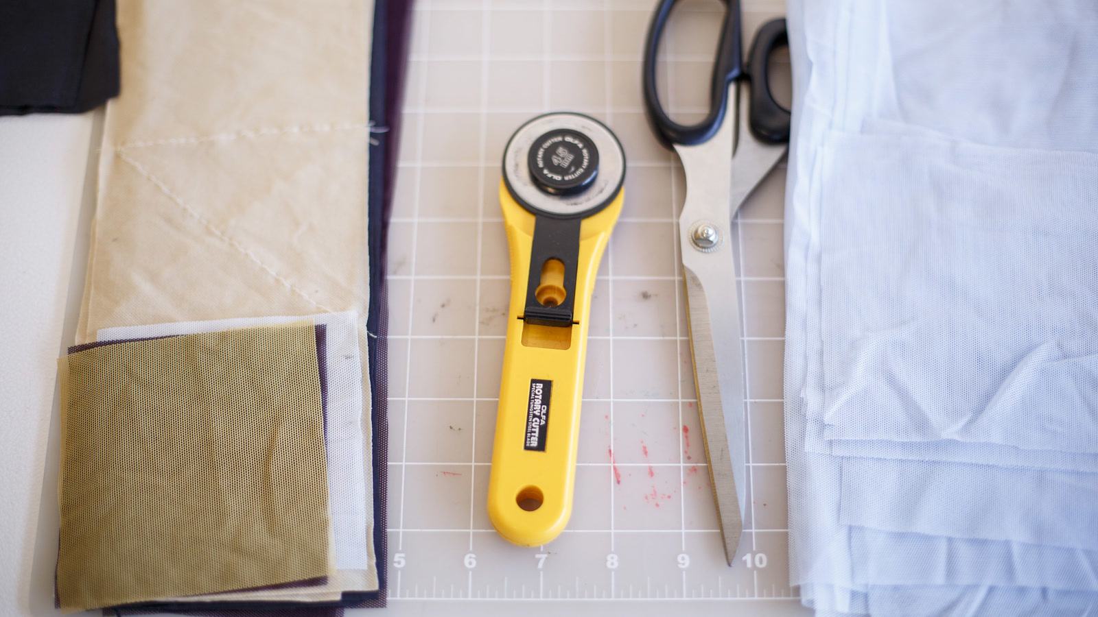 Textile-development table with layered mesh specimens, shears, a rotary cutter, measuring grid, and pale fabric